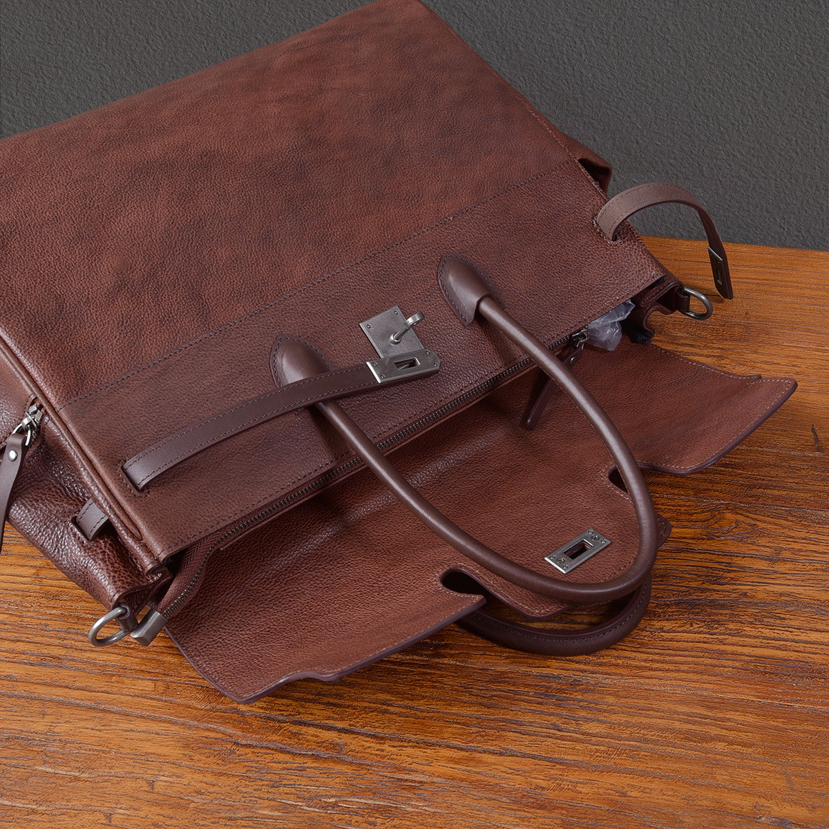 Brown leather work bag with dual handles and zipper, designed for business use, displayed on a wooden surface.