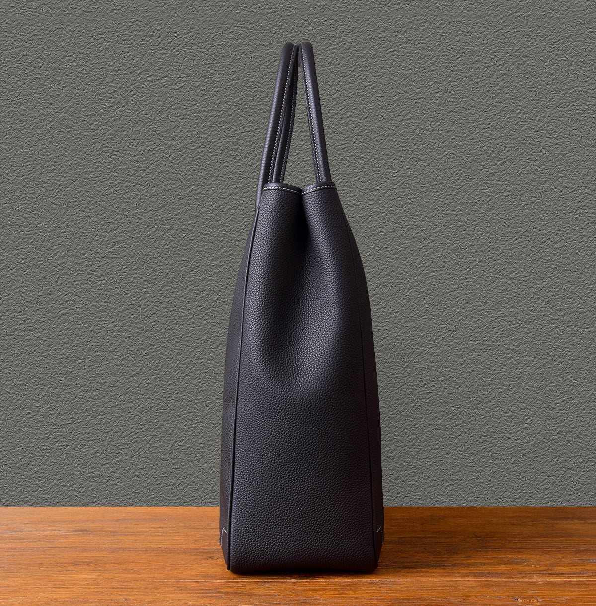 Large vertical cowhide tote bag in black leather, showcasing its sleek side profile on a wooden surface.