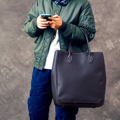 Person in a green bomber jacket holding a smartphone and carrying a large black leather tote bag.