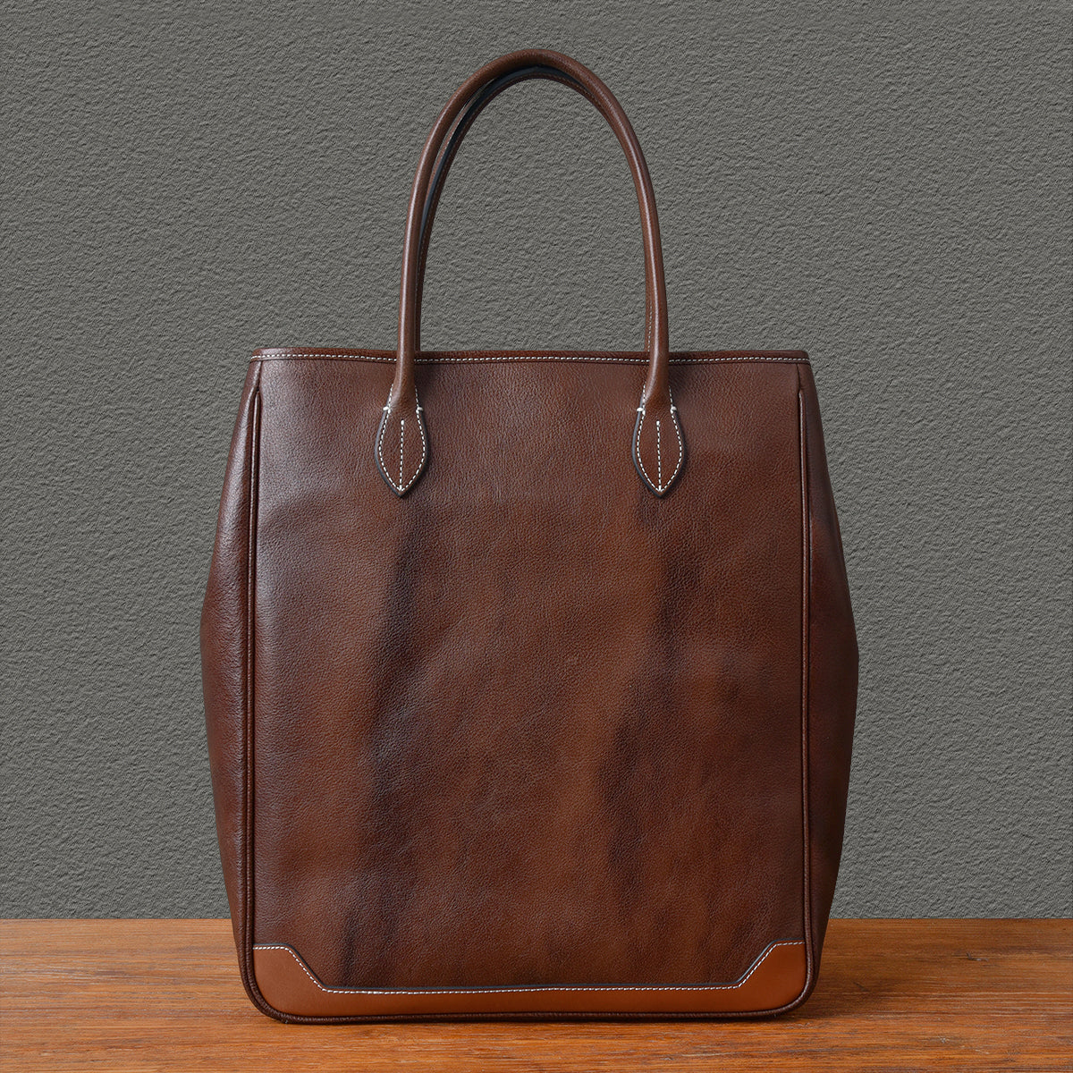 Large vertical brown leather tote bag with sturdy handles and minimalist design.