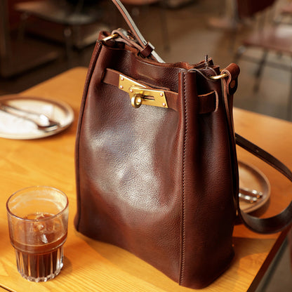 Italian Leather Bucket Bag Women in brown color, featuring a gold clasp, placed beside a glass of drink on a wooden table.