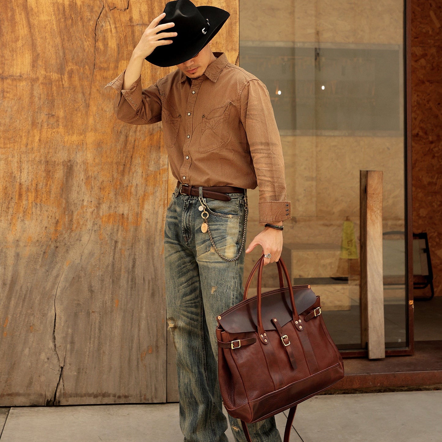 Man wearing a brown shirt and hat, holding an Italian Leather Travel Bag, standing by a rustic backdrop.